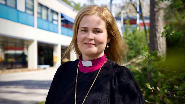 Piispa Mari Leppänen seisoo rakennuksen edessä ja katsoo kohti kameraa. 