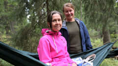 Sari Kalenius ja Tiina Martikainen hymyilevät riippukeinussa metsässä.