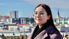 Silmälasipäinen nuori tummatukkainen nainen katsoo kameraan taustallaan Tallinnan kaupunki.