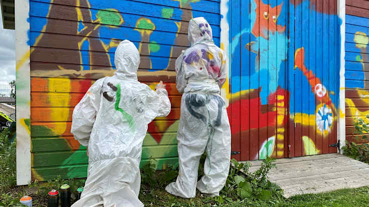 Valkoisiin suojahaalareihin pukeutuneet lapset spraymaalaavat ladon seinää. Maassa on spraymaalipurkkeja. 
