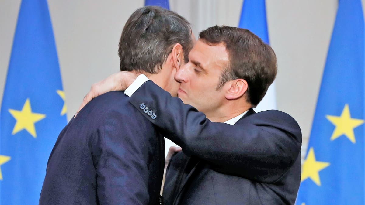 Mitsotakis ja Macron halaavat. Takana näkyy EU-lippuja.