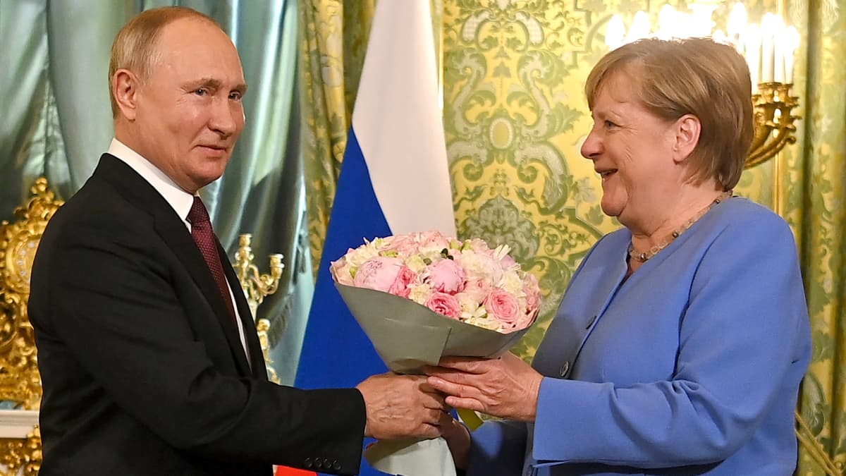 Venäjän presidentti Vladimir Putin antaa ruusukimpun Saksan liittokansleri Angela Merkelille, taustalla koristeellista tapettia ja Venäjän lippu.