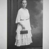 Nainen poseeraa mustavalkoisessa valokuvassa vakava ilme kasvoillaan. Yllä vaalea pitkä mekko, kädessä musta käsilaukku.
