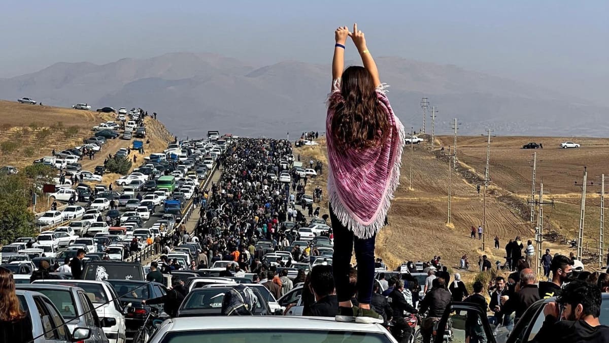 Mielenosoittaja seisoo auton katolla kädet pystyssä. Taustalla suuri väkijoukko seisoo tien päällä.