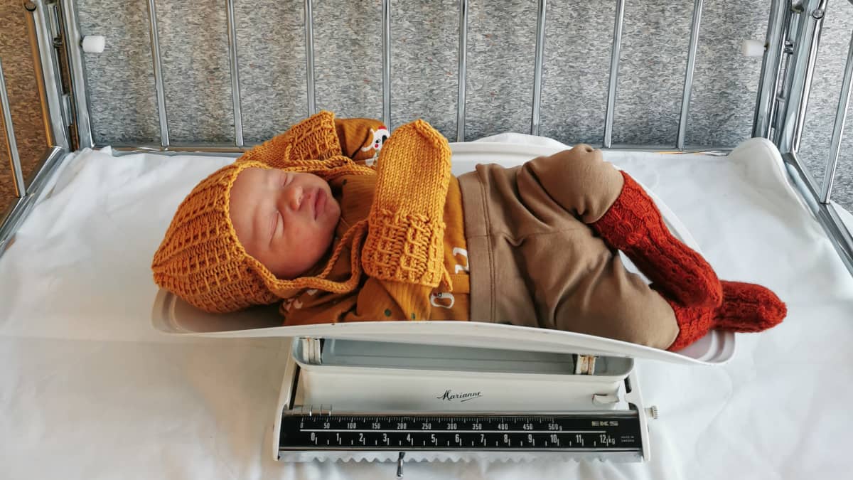 Vauva vaa'alla vaatteet päällä. Vauvan yllä taulut, joissa on merkittynä: 3.12.2021 4:47 Ramppi 71 Volvo XC90 3,750 g 49 cm