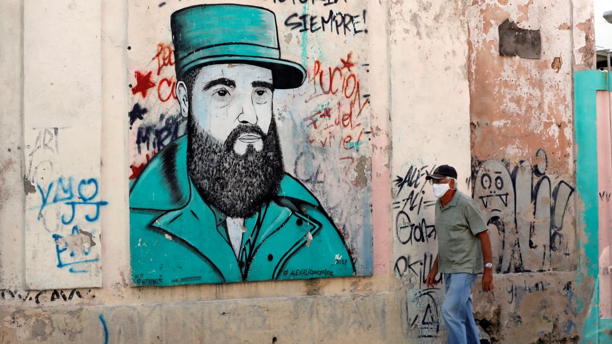 Valkohiuksinen mies mustassa lippalakissa kävelee kadulla. Seinään on maalattu naivistinen kuva parrakkaasta Fidel Castrosta. Sen yllä lukee: "Hasta la victoria siempre!"