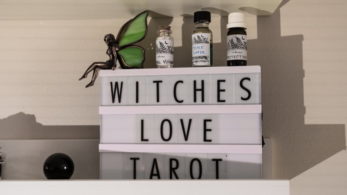Neliön muotoinen pöytävalo, jossa on teksti "witches love Tarot". Lampun päällä on öljyjä ja keijukoriste.