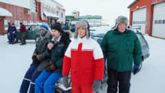 Talvivaatteisiin pukeutuneita miehiä teollisuuslaitoksen pihalla. Kaksi miestä seisoo, kaksi istuu lumisen auton keulan päällä. Ilmeet ovat hymyttömät.
