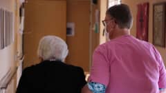 En kvinna i vårduniform och munskydd håller en äldre dam i armen.