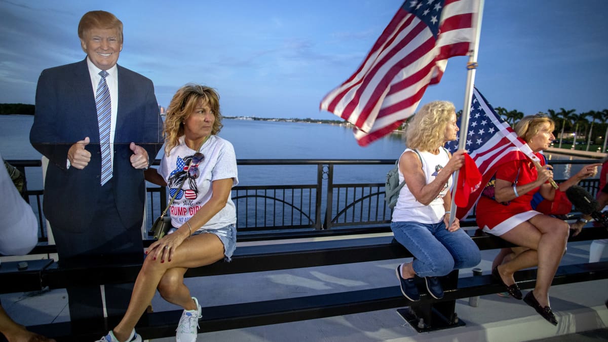 Trumpin kannattajia iltavalossa istuu sillalla. Yhdellä on Trump pahvikuva ja kahdella muulla USA:n lippu. 