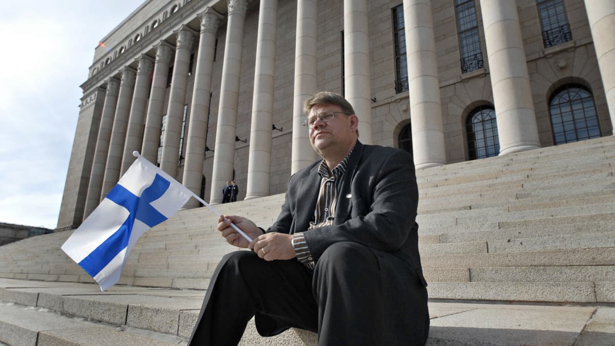 Timo Soini istuu eduskuntatalon portailla Suomen lippu kädessään. 