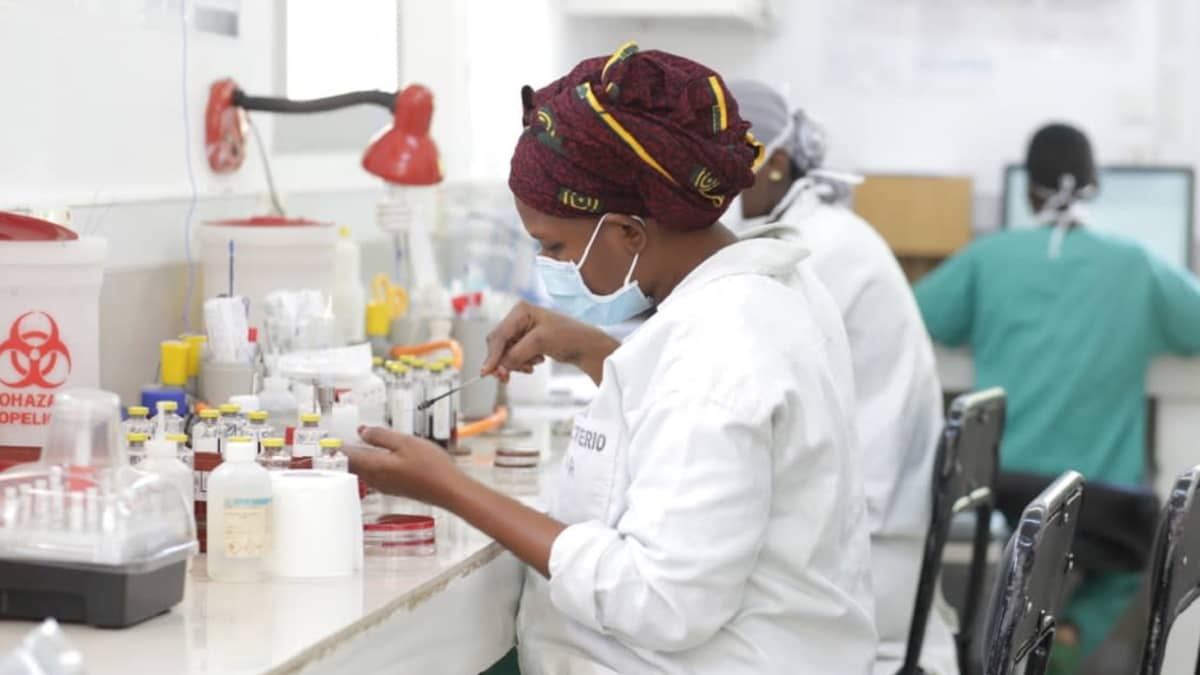 Maskiin, valkoiseen takkiin ja ruskeaan kuviolliseen huiviin pukeutunut nainen tutkii näytettä laboratoriopöydän ääressä.