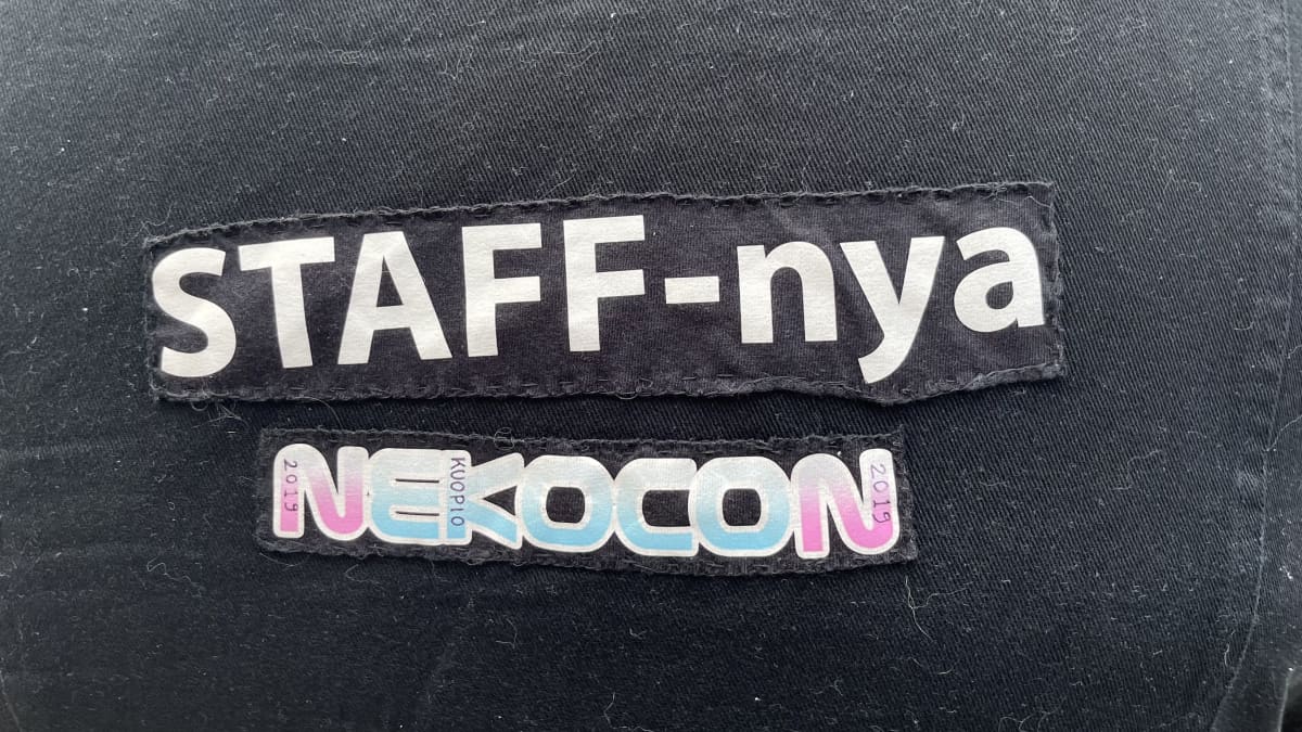 Mustan farkkuliivin selkämyksessä kangasmerkit, joissa yhdessä lukee "staff-nya" ja toisessa "nekocon".