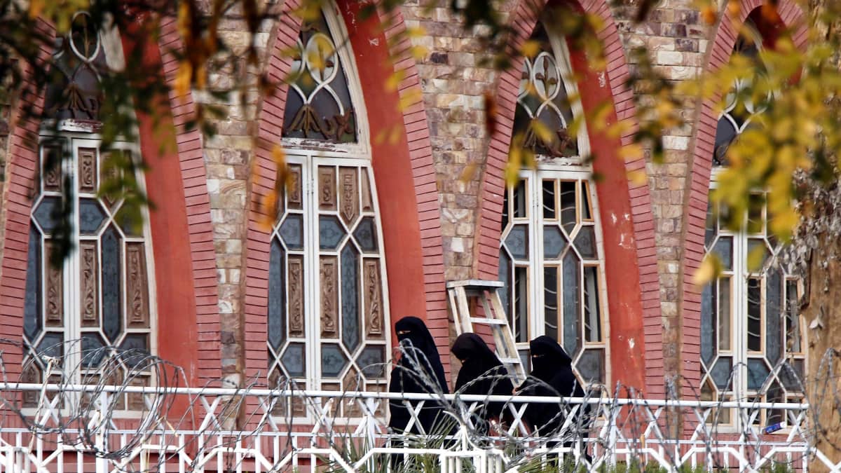 Kolme mustiin koko vartalon ja kasvot peittäviin asuihin pukeutunutta naista kulkee valkoisen aidan takana punatiilisen rakennuksen seinäniertä suurten ovien editse.