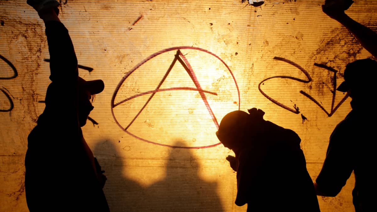 Kolme tummaa hahmoa seinään maalatun anarkistien tunnuksen edessä.