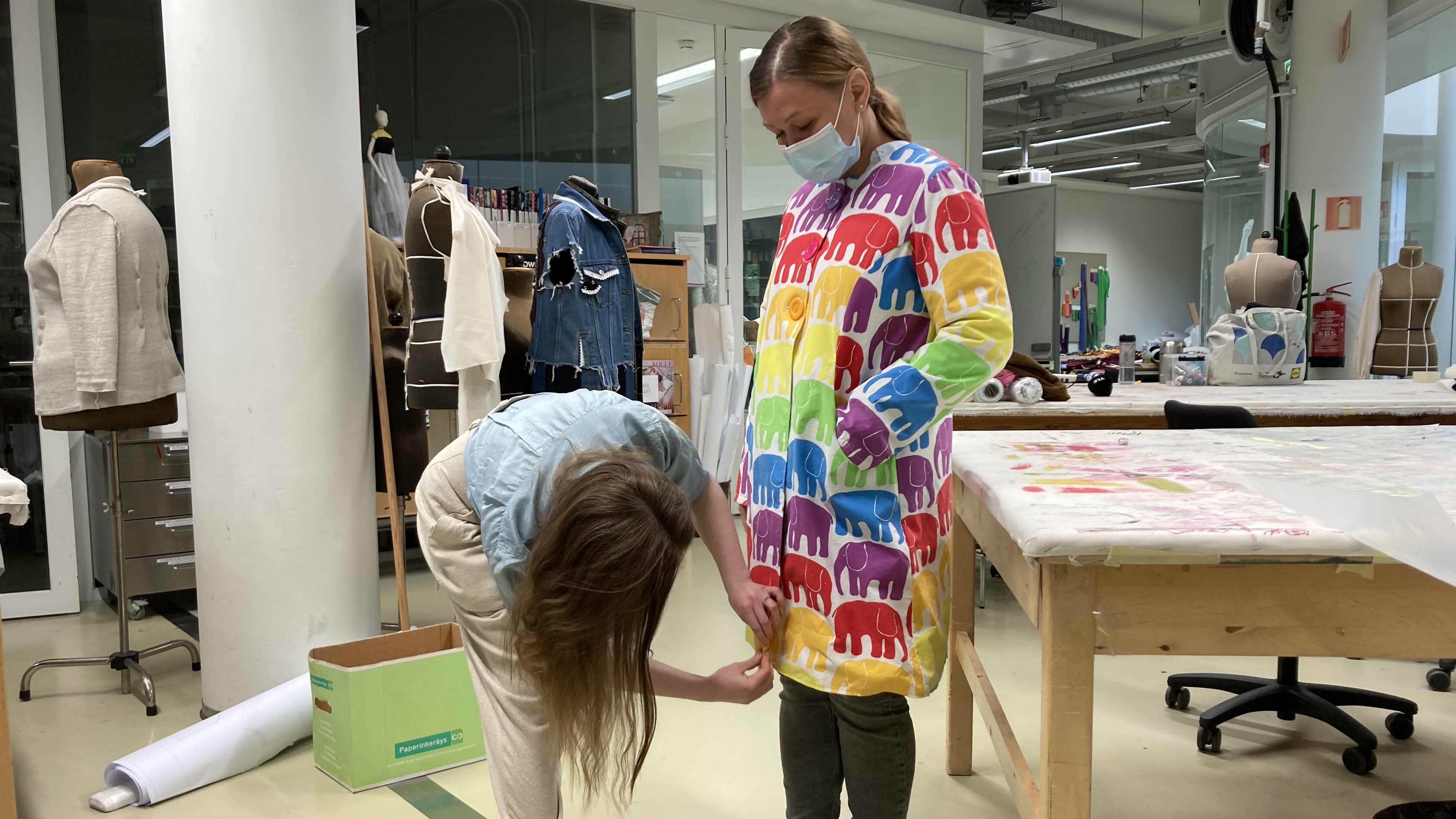 Opettaja Iida Silvennoinen asettelee elefanttikuvioista takkia, joka on muoti- ja puvustusalan opiskelija Piia Selinkosken päällä.