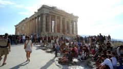 Turistiryhmiä istumassa, seisomassa ja kävelemässä. Taustalla näkyvät Parthenonin temppelin rauniot auringonpaahdetta vasten.