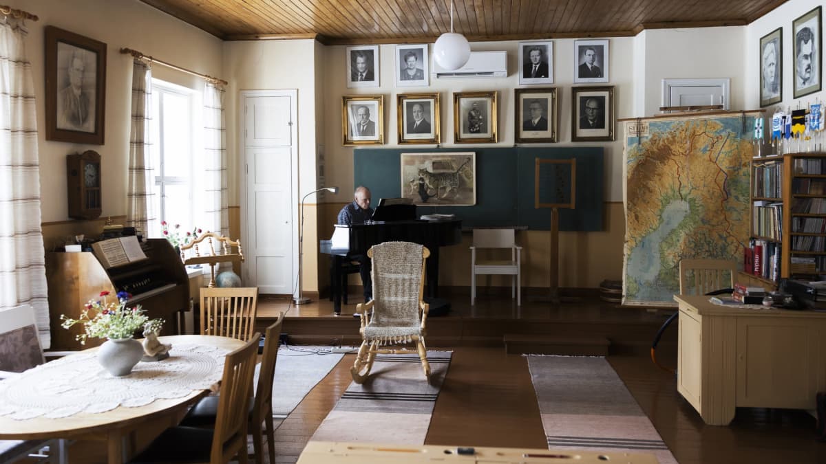 Vanha mies pianon ääressä. Seinillä presidenttien kuvia ja vanha Suomen kartta. Räsymatot sekä keinutuoli. Harmooni ja kukkia pöydällä.