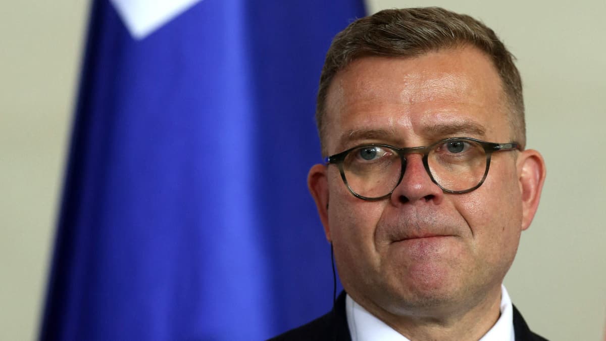 Petteri Orpo katsoo ohi kamerasta huulet tiiviisti yhdessä. Taustalla on Suomen lippu. Orpolla on pyöreät silmälasit, puku ja siniraidallinen kravatti.