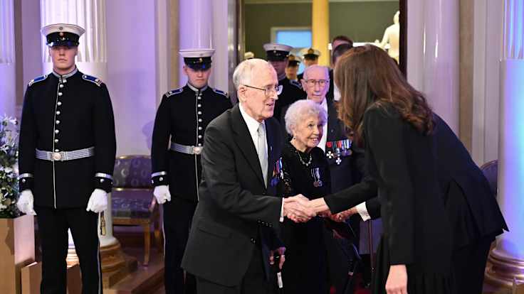 A woman with long brown hair, wearing a dark suit seen from the side, shaking hands with an elderly man, with uniformed soldiers in the background.