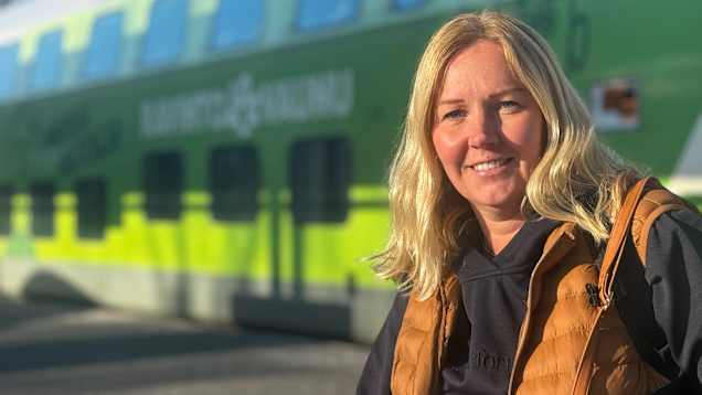 Mia Nyholm framför ett grönt och vitt tåg, klädd i en brun väst och svart tröja. Det är soligt och skuggan faller på marken bakom personen.