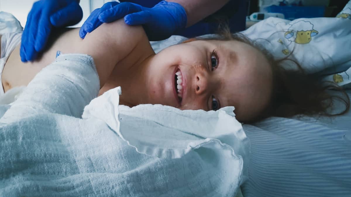 Hilda Tolonen nauraa sairaalasängyssä kun kumihanskoihin verhoutuneet kädet koskettavat hänen selkäänsä.