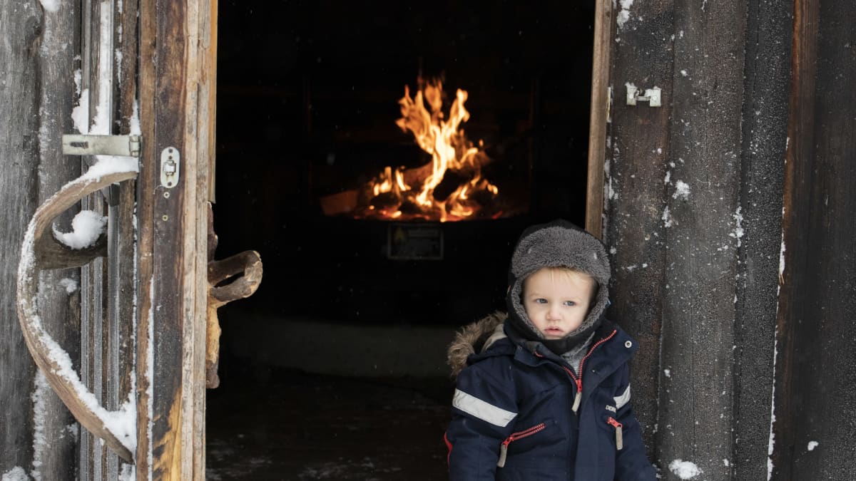 Pieni poika seisoo talvivarusteissa kodan edessä, kodassa palaa nuotio.