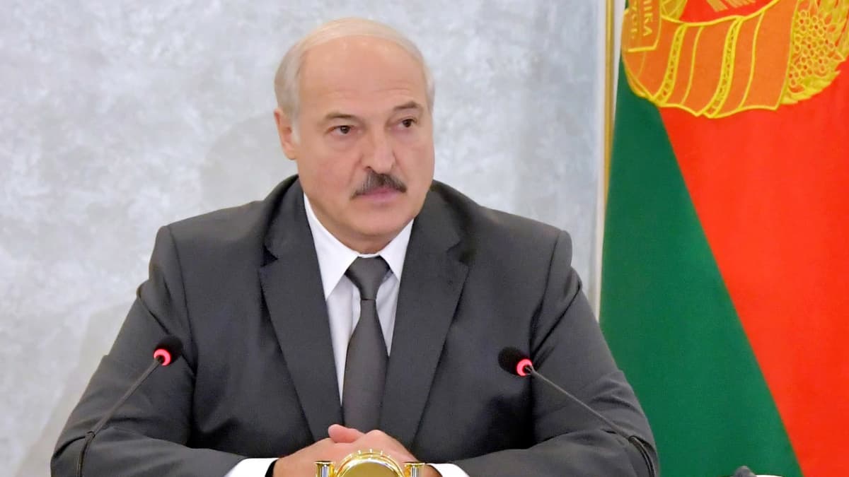 Lukashenka istuu pöydän takana tummanharmaassa puvussa ja kravatissa, Hänen edessään on mikrofoni. Hän on ristinyt kätensä eteensä, Taustalla näkyy Valko-Venäjän punavihreä lippu.