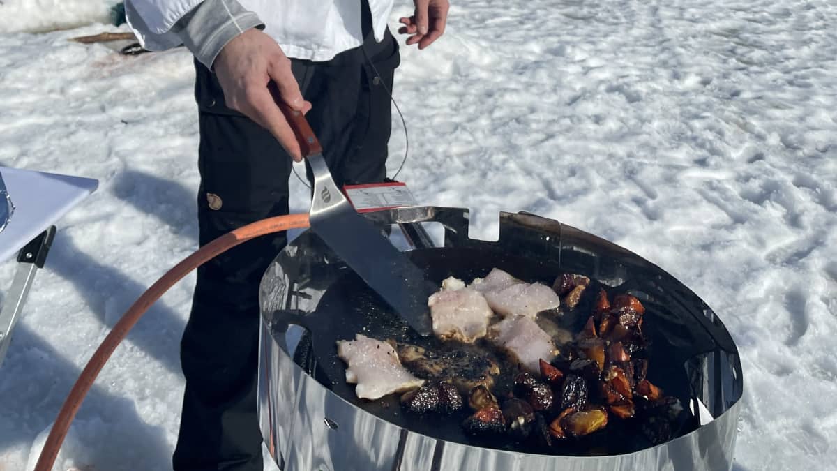 Mies kokkaa ruokaa kenttäkeittiössä järven jäällä, päällään kokkiasu.