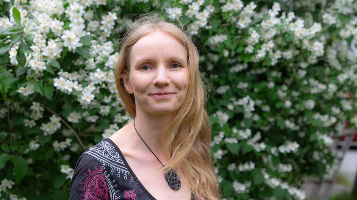 Kesämekkoon pukeutunut tietokirjailija Marika Haataja katsoo suoraan kameraan ja hymyilee. Taustalla näkyy rehevä pensas, jossa on paljon valkoisia kukkia.