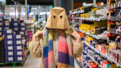 Nainen seisoo supermarketin makeisosastolla ruskea paperikassi päässään. Kassiin on leikattu silmän reiät.