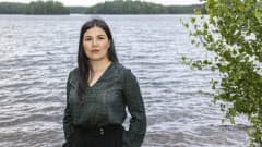 Mustatukkainen nainen seisoo koivun vieressä järven rannalla.