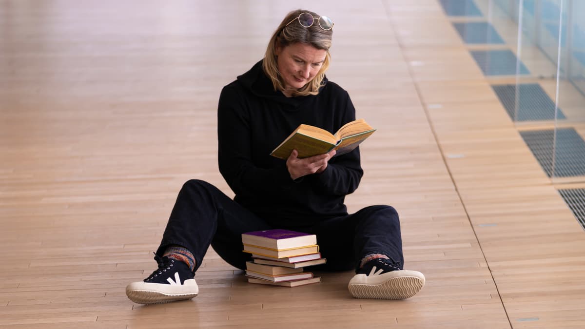 Johanna Kaakinen istuu Oodin lattialla kirjapinon kera ja lukee kirjaa.