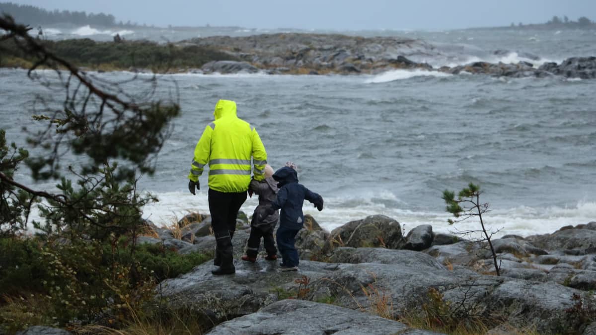 En pappa med två barn går på klippor i hård vind. Vattnet slår mot klipporna.