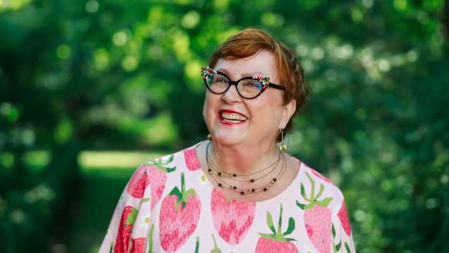 En skrattande person med en färgglad tröja med jordgubbsmönster står utomhus omgiven av grönska.