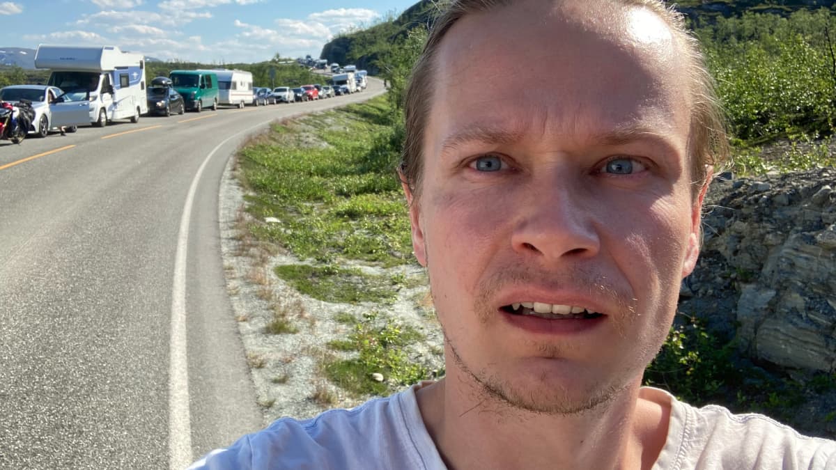 Janne Kulju katsoo kameraan Kilpisjärven rajalla, Norjan puolella. Taustalla pitkä autojono.