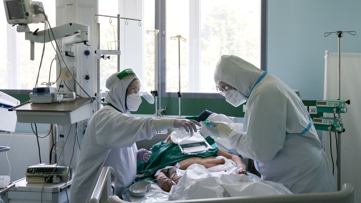 Kaksi suojapukuihin pukeutunutta sairaalatyöntekijää, välissä koronapotilas sängyllä.