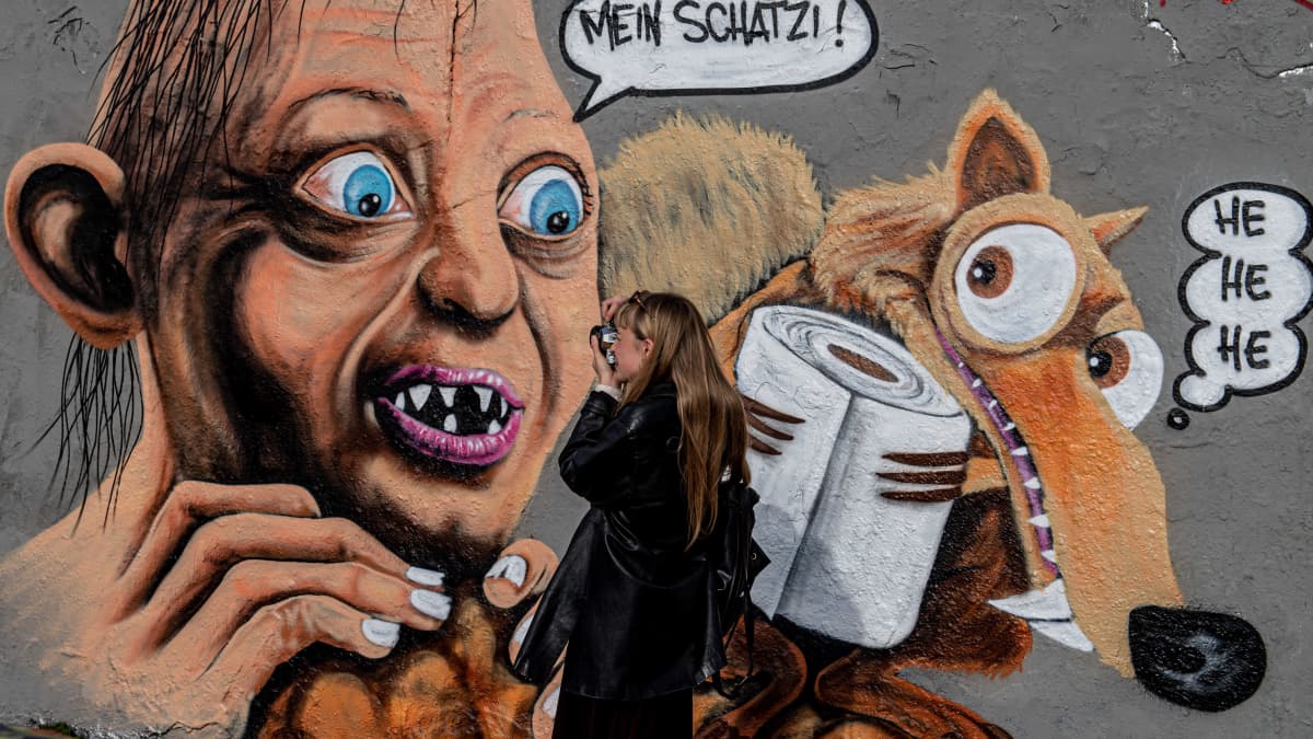 Nainen ottaa kuvaa Berliinin entiseen muuriin maalatun graffitin edustalla. Kuvassa Taru sormusten herrasta -trilogiasta tuttu Klonkku-hahmo on löytänyt uuden "aarteen" koronakriisin hengessä.