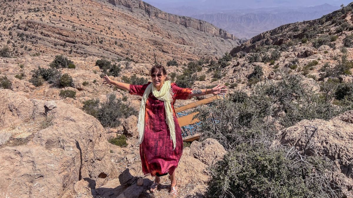 Nainen poseeraa käsiään levittäen kameralle punaisessa mekossa ja vaaleassa huivissa. Taustalla vuoristoa.