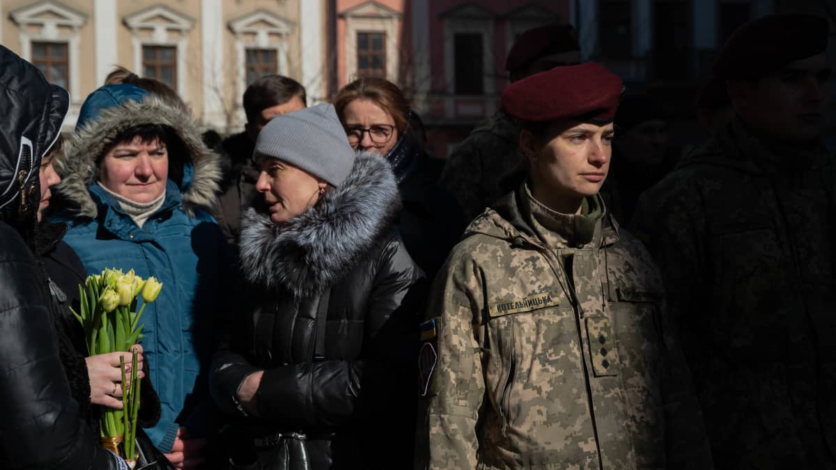 Oikealla ukrainalainen naissotilas ja vasemmalla ukrainalaisia siviilejä osallistumassa hautajaisiin. 