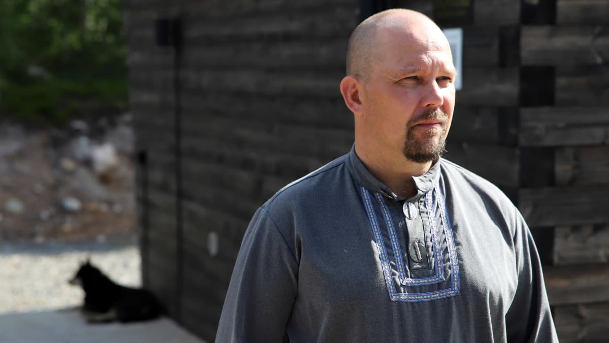 Mies seisoo harmaassa paidassa harmaata aittaa vasten, taustalla näkyy koira