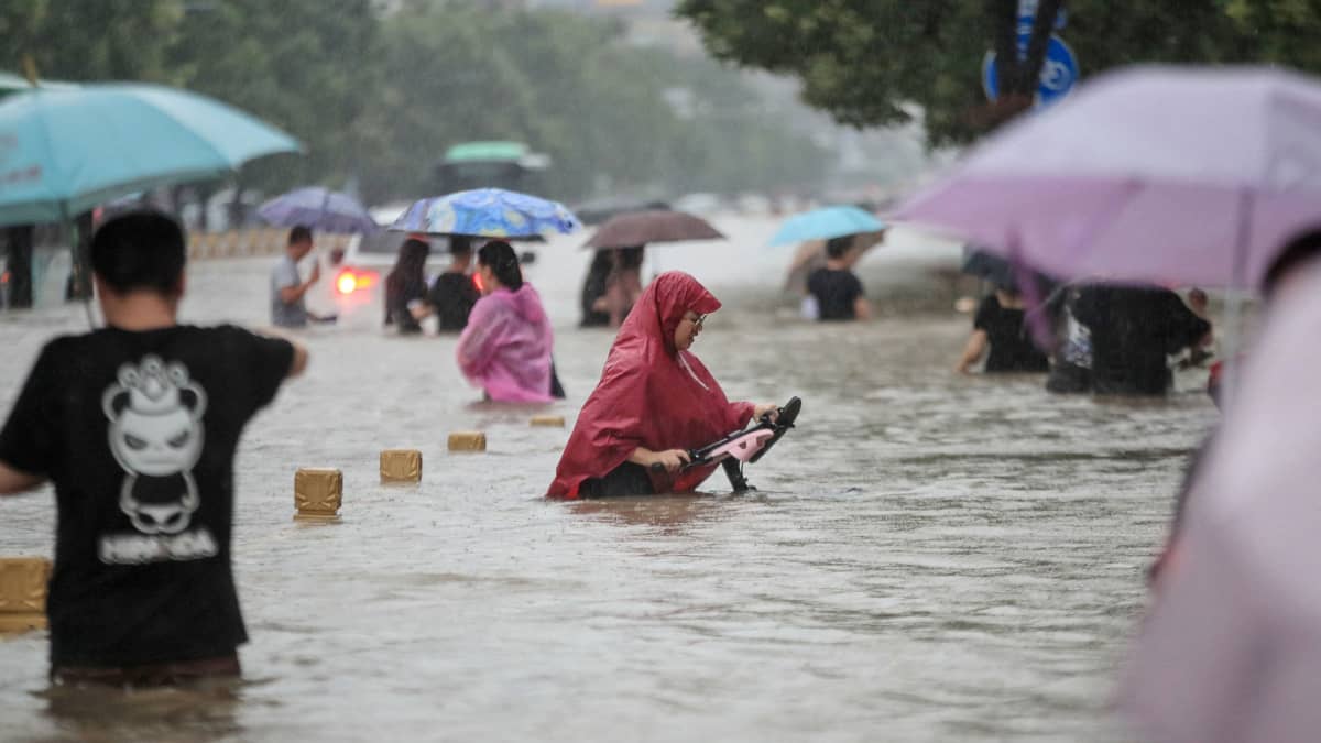 Ihmiset kävelevät tulvivalla kadulla sateenvarjojen ja sadeasujen kanssa. 
