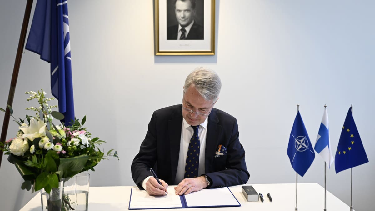 Pukuun pukeutunut henkilö (Pekka Haavisto) kirjoitaa kynällä paperiin. Pöydällä valkoinen pöytäliina, kukkakimppu ja kolme pientä lippua; Nato, Suomi, EU..