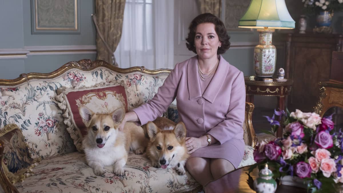 Olivia Colman som spelar drottning Elizabeth sitter i en antik soffa i en vacker salong med två av sina Corgis intill sig.