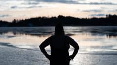 Nainen uimapukusillaan seisoo silhuettina uimarannalla katsoen puoliksi jäätynyttä lahtea.