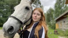 Kuvassa poseeraa hevosen kanssa  Kuhmoisten ratsastuslukiota käyvä 18-vuotias Silja Lehtonen. Hevosen pää on Siljan poskea vasten. Hevonen on vaalea. 