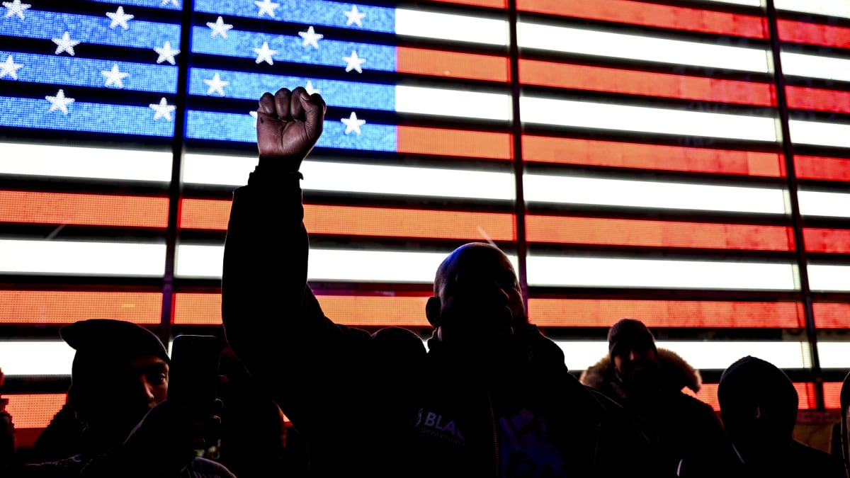 Etualalla miehiä, joista yksi nostaa nyrkkiään pystyyn, ja tautalla Yhdysvaltain lippu näytöllä.
