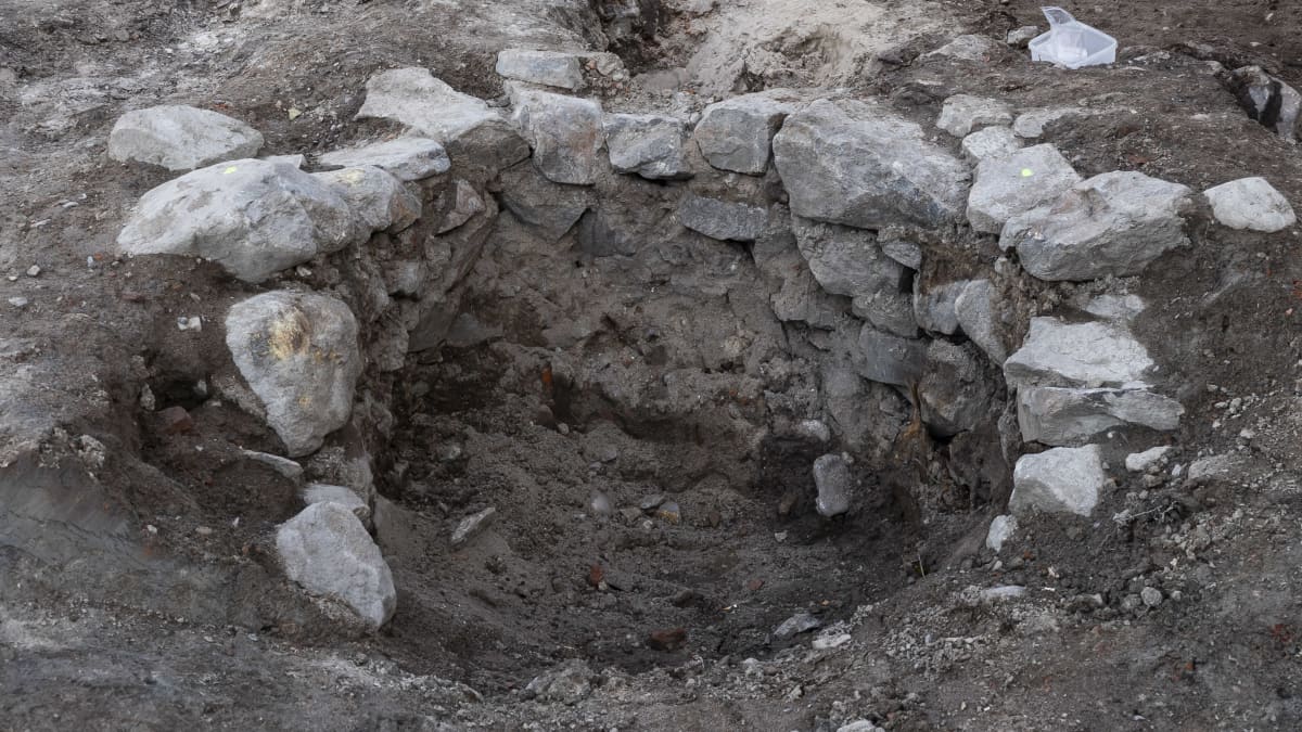 Etualalla kuvassa on kaivo. Taaempana kolme arkeologia suorittamassa kaivauksia. 