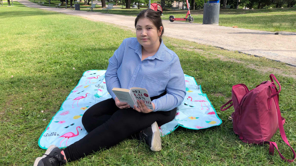 Tummahiuksinen nainen istuu viltillä nurmikolla lukemassa kirjaa. 