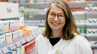 En kvinnlig apotekare i vit rock står i sitt apotek och ler mot kameran. 